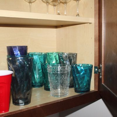 Sale Photo Thumbnail #3: Collection of colorful acrylic and glass drinking tumblers arranged in a kitchen cabinet alongside stemmed wine glasses on the shelf above.