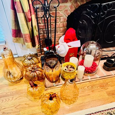 Sale Photo Thumbnail #21: Collection of seasonal decor, featuring amber glass pumpkins and gourds, a glass owl pitcher, pillar candles, and a cloche display set near a fireplace.