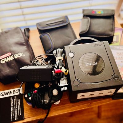 Sale Photo Thumbnail #71: Black Nintendo GameCube console bundle with controller, cables, Super Smash Bros. Melee game, and vintage Game Boy carrying cases.