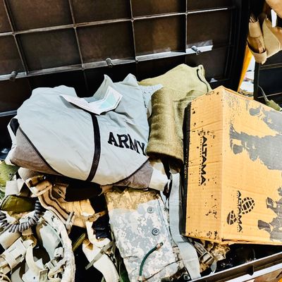 Sale Photo Thumbnail #27: A plastic storage trunk filled with military gear, including a grey US Army jacket, digital camouflage items, tactical helmet accessories, and a cardboard Altama shoe box.