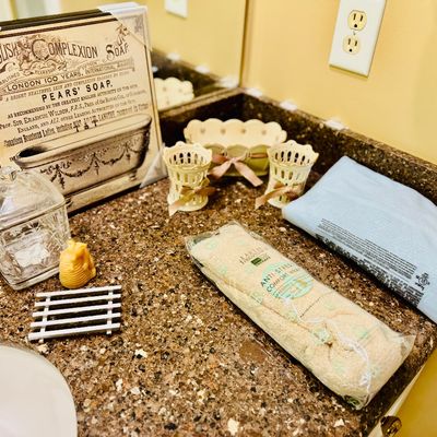 Sale Photo Thumbnail #68: Collection of bathroom counter decor and accessories, including a framed vintage-style Pears' Soap advertisement, etched glass apothecary jar, ceramic baskets with bow accents, and wrapped spa towels.