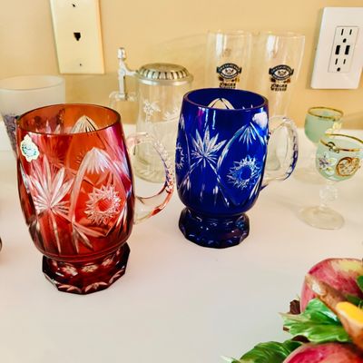 Sale Photo Thumbnail #37: Pair of cut-to-clear crystal mugs in vibrant red and cobalt blue with detailed starburst and pinwheel motifs, displayed alongside other glassware.