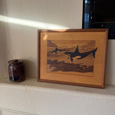 Framed wood marquetry artwork depicting swimming orca whales, shown alongside a small studio pottery jar with a lid.