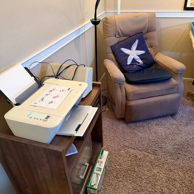 White HP DeskJet printer sitting atop a wooden side table alongside a tan upholstered recliner chair with a starfish throw pillow.