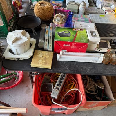 Folding table laden with various household goods, including a ball of twine, cast iron skillet, boxed Coleman air pump, lighting...