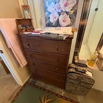 Dark wood five-drawer chest of drawers topped with a floral canvas painting, small decorative items, and packaged linens, accompanied by...
