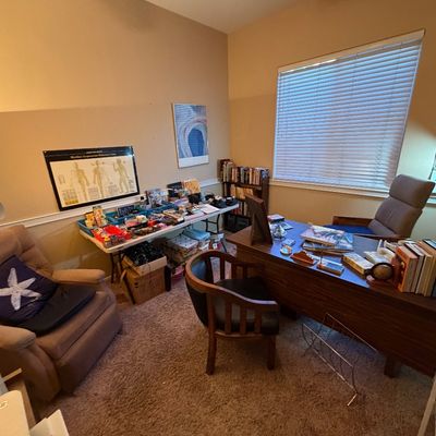 Home office setup featuring a wooden executive desk, upholstered armchairs, folding tables with miscellaneous sale items, and anatomical...