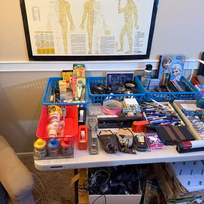 A folding table displaying assorted office supplies, stationery, and small tools, with a laminated Touch for Health Meridian Acupuncture...