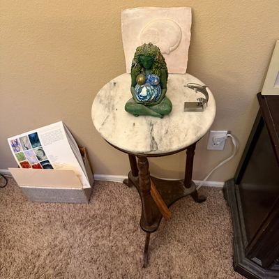 A round marble-top plant stand displaying a Mother Earth Gaia figurine and a small silver dolphin sculpture.