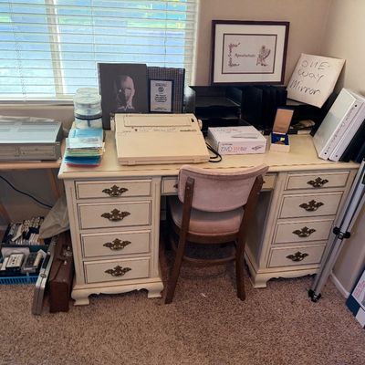 Vintage white pedestal desk with brass handles and matching chair, featuring various office supplies, media electronics, and framed artwork.