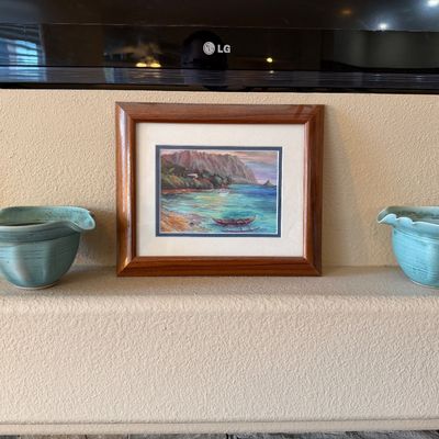 Framed coastal landscape painting featuring an outrigger canoe, flanked by a pair of light blue glazed studio pottery bowls.