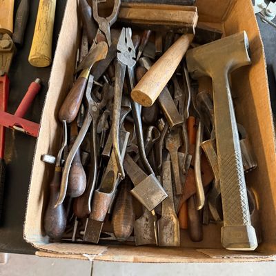 Cardboard box filled with assorted vintage hand tools, including pliers, chisels, awls, and a brass mallet.