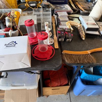 Assorted household items, hand tools, hummingbird feeders, light bulbs, and workshop accessories arranged on folding tables.