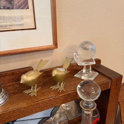 Pair of brass bird-shaped tea light candle holders displayed on a wooden shelf alongside a crystal paperweight and decanter set.