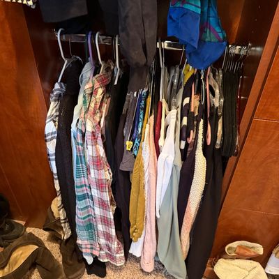 Selection of women's casual clothing hanging on plastic and wire hangers in a wooden closet frame, alongside various boots below.