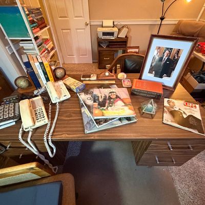 Vintage wood grain executive office desk cluttered with items including landline telephones, art and history books, magazines, small...