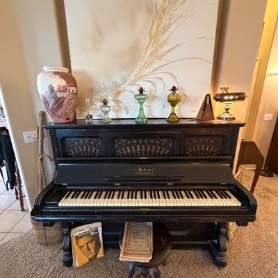 Vintage Estey dark wood upright piano paired with a piano stool, surrounded by home decor including oil lamps, a large canvas painting,...
