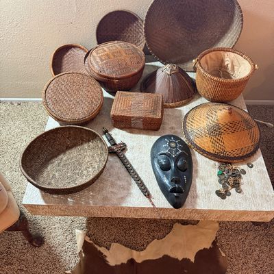 Collection of woven Asian and African style baskets, straw hats, a carved black mask, and decorative coin crafts displayed on a coffee...