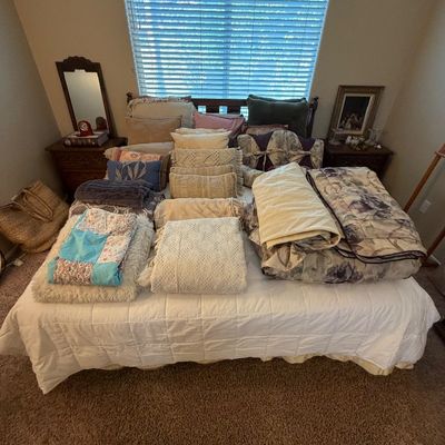 Large collection of bedding, decorative throw pillows, comforters, and quilts neatly arranged on a bed, flanked by wooden nightstands...