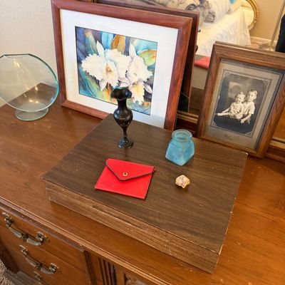 Collection of tabletop decor including framed floral artwork, a vintage black and white portrait, a small brass vase, a red pouch, and...