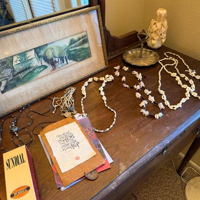 Assortment of shell necklaces, costume jewelry, and decor displayed on a vintage wooden dresser, featuring a framed landscape print and a...