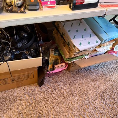 Collection of office supplies, electronics, headphones, and assorted cables stored in cardboard boxes beneath a folding table.
