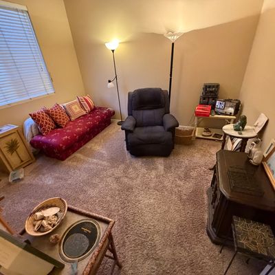 A furnished room with a red pattern-upholstered daybed couch, a blue plush recliner, floor lamps, small end tables, and framed artwork.