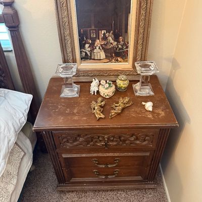 Traditional wooden nightstand with carved details, topped with a framed art print of Las Meninas, clear glass candlesticks, and small...