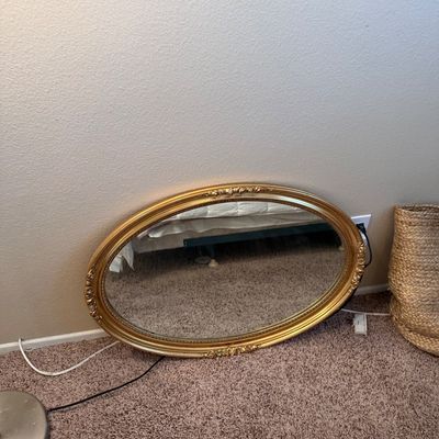 Vintage oval wall mirror with an ornate gilt frame and beaded trim resting against a wall.