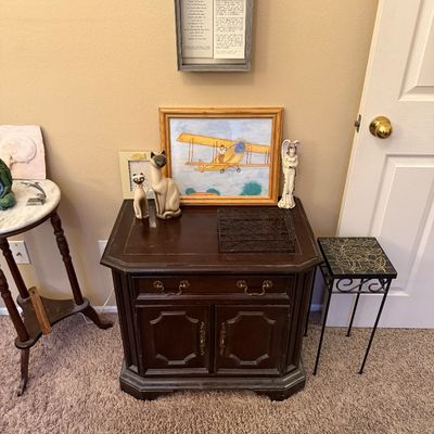 Dark wood accent cabinet topped with ceramic Siamese cat figurines, framed artwork of a biplane, an angel statue, and framed wall art above.