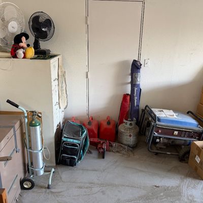 Assorted household equipment including metal filing cabinets, a portable generator, gas cans, a hand-truck-mounted tank, and electric fans.