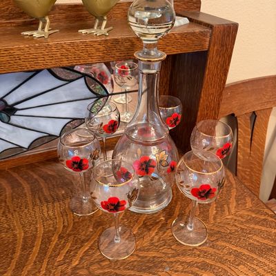 Vintage glass liquor decanter set featuring painted red flowers and gold accents, accompanied by matching stemware glasses.