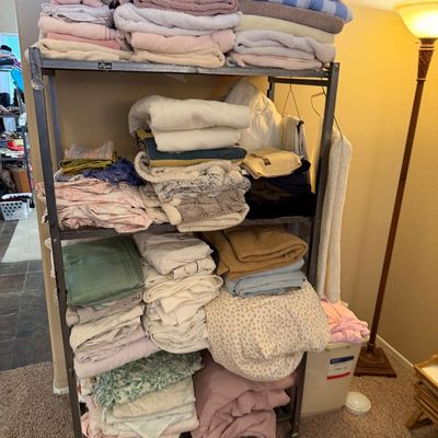 Four-tier metal rolling shelving unit stacked with various bath towels, bed sheets, and household linens.