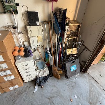 Assortment of garage items including cleaning tools, brooms, a step ladder, a rolling crate, hand weights, and a small metal filing cabinet.