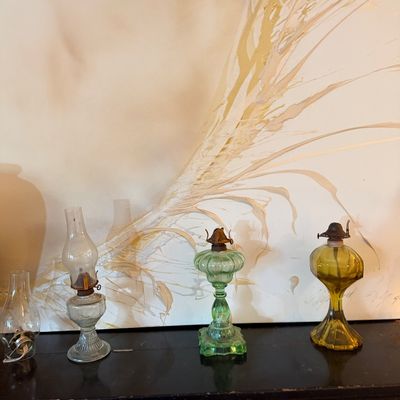 Collection of vintage glass oil lamps, including clear, green pressed glass, and amber glass bases, displayed in front of a textured...