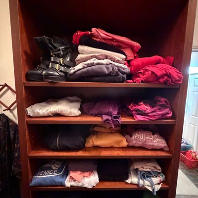 Tall dark wood bookcase filled with stacks of folded casual clothing and black buckle boots on the top shelf.
