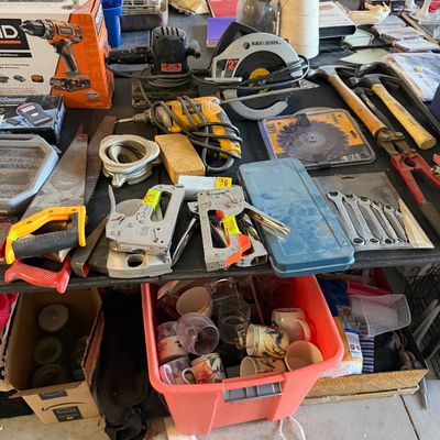 A wide assortment of workshop power and hand tools displayed on a table, including a Black & Decker circular saw, DeWalt corded...