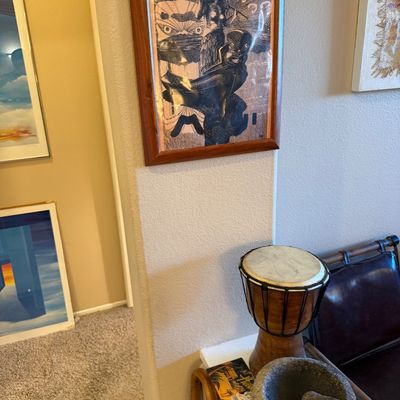 Framed Polynesian-style artwork hanging on a wall, shown alongside a djembe drum and a stone molcajete with pestle.