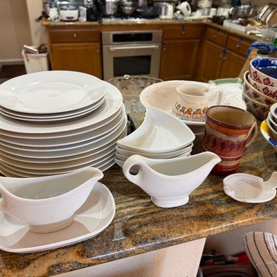 Collection of white ceramic dinnerware, gravy boats, crescent side dishes, and pottery mugs displayed on a granite countertop.