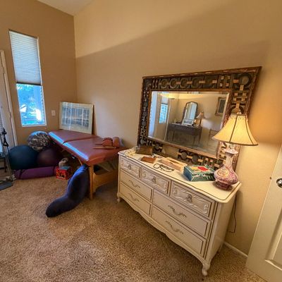 Off-white French Provincial style double dresser with an ornate gold frame mirror, table lamp, and portable massage table in a bedroom.