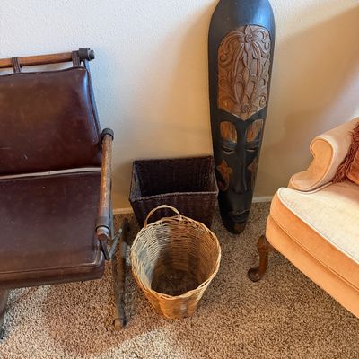 Tall hand-carved wooden African-style wall mask standing beside woven storage baskets, an antique leather armchair, and a velvet...