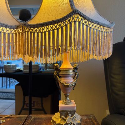 Vintage Hollywood Regency style table lamp featuring a pink marble urn-form base with brass cherub accents and an ornate fabric shade...