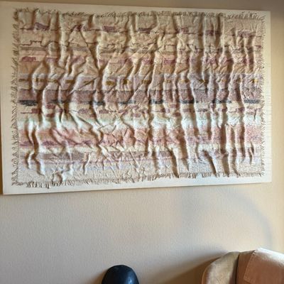 Large mounted woven fiber art piece featuring neutral and pastel tones with heavy texture and fringe details.