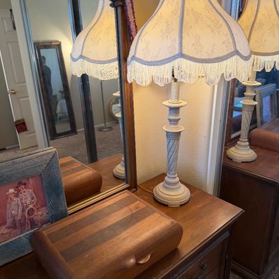 Vintage white painted table lamp with a scalloped, fringe-trimmed fabric shade sitting on a wooden dresser beside a striped wooden...