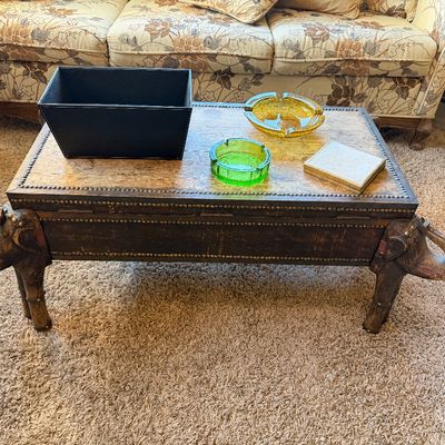 Vintage carved wooden elephant coffee table with brass nailhead trim detail, topped with mid-century glass ashtrays and decorative...