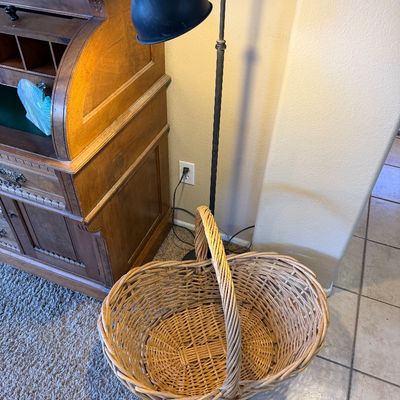 Large woven wicker basket with an arched handle, displayed alongside a black metal floor lamp and a vintage wooden roll-top desk.