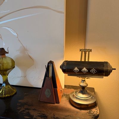 Vintage brass piano lamp with a perforated cylinder shade, displayed alongside a wooden metronome and a green glass oil lamp.