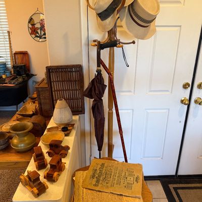 Wooden coat rack holding straw fedora hats, a brown umbrella, and a walking cane, alongside a small side table displayed with vintage...