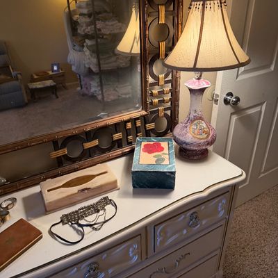 French Provincial style white dresser featuring a large ornate wall mirror and a porcelain table lamp with an embroidered shade.
