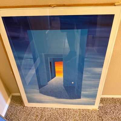 Framed architectural surrealist art print depicting a blue corridor opening onto an orange sunset sky, mounted under glass in a simple...